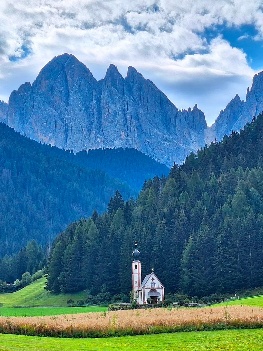 A small white chapel below jagged mountain peaks, with meadows and forest