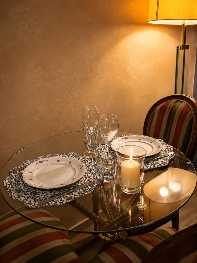 Candle-lit glass table set for two beside a warm floor lamp